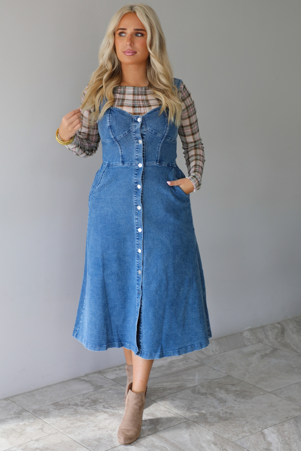 A woman wearing a medium wash denim midi dress with button closures along the  front, with pockets on either side, paired with a sheer, multi-colored plaid long sleeved top underneath, standing indoors against a white wall with white marble flooring. 