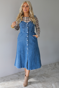 A woman wearing a medium wash denim midi dress with button closures along the  front, with pockets on either side, paired with a sheer, multi-colored plaid long sleeved top underneath, standing indoors against a white wall with white marble flooring. 