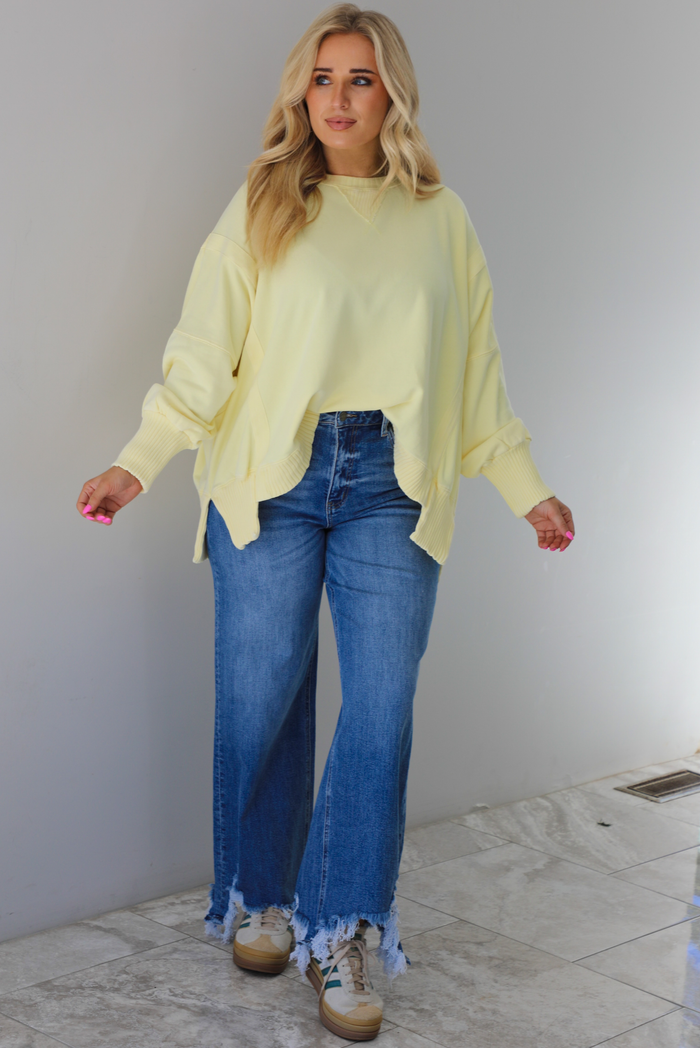 Woman wearing a pale yellow oversized sweater with dark wash denim and sneakers. The photo is taken indoors against a white wall with white marble flooring. 