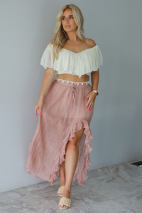 Woman wearing a cream cropped, off the shoulder top with a high waisted blush maxi skirt indoors against a white wall with white marble flooring. 