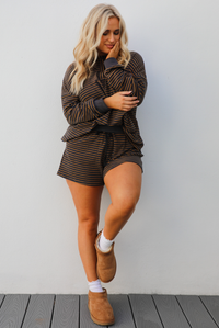 Woman model wearing a brown and multi-colored striped pattern throughout. The set consists of an oversized top with quarter-button details and shorts. The photo is taken outdoors against a white wall. 