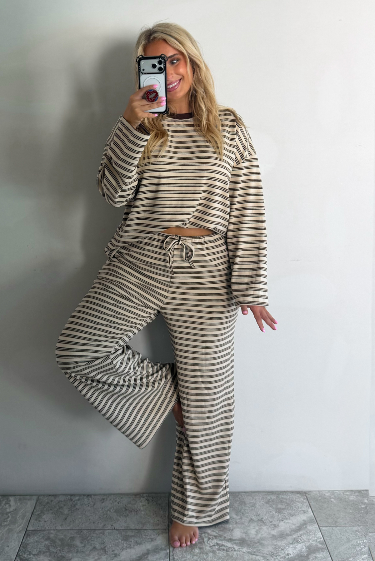 A woman wearing a sand colored matching two piece set with a brown striped pattern throughout featuring an oversized long sleeved top and oversized slouchy wide leg pants, standing indoors against a white wall with white marble flooring taking a mirror selfie. 