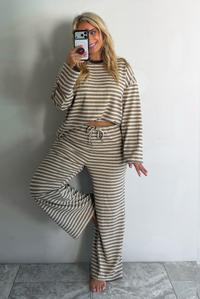A woman wearing a sand colored matching two piece set with a brown striped pattern throughout featuring an oversized long sleeved top and oversized slouchy wide leg pants, standing indoors against a white wall with white marble flooring taking a mirror selfie. 