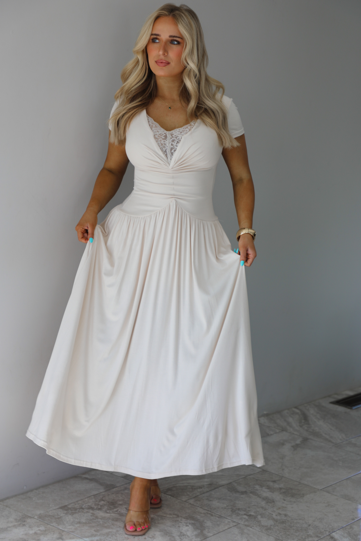 Woman wearing a light sand colored maxi length dress that has a fitted bodice with lace detailing in the center of the bust, a drop waist design, short sleeves, and a flowy skirt. The photo is taken indoors against a white wall with white marble flooring. 
