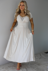 Woman wearing a light sand colored maxi length dress that has a fitted bodice with lace detailing in the center of the bust, a drop waist design, short sleeves, and a flowy skirt. The photo is taken indoors against a white wall with white marble flooring. 