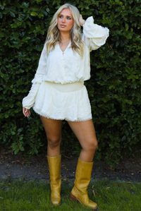 Woman model wearing an off-white romper with yellow boots outdoors in front of greenery. 