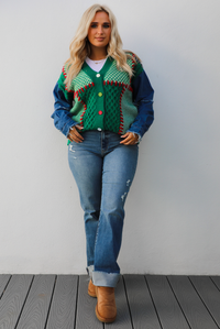 Woman model wearing a green and multi-colored cardigan with denim sleeves, medium wash denim jeans, and tan uggs. The photo is taken against a white wall outdoors.