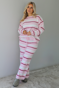 Woman wearing a pink set with a darker pink and white striped design. The set consists of an oversized sweater top and high waisted pants. The photo is taken indoors against a white wall with white marble flooring. 