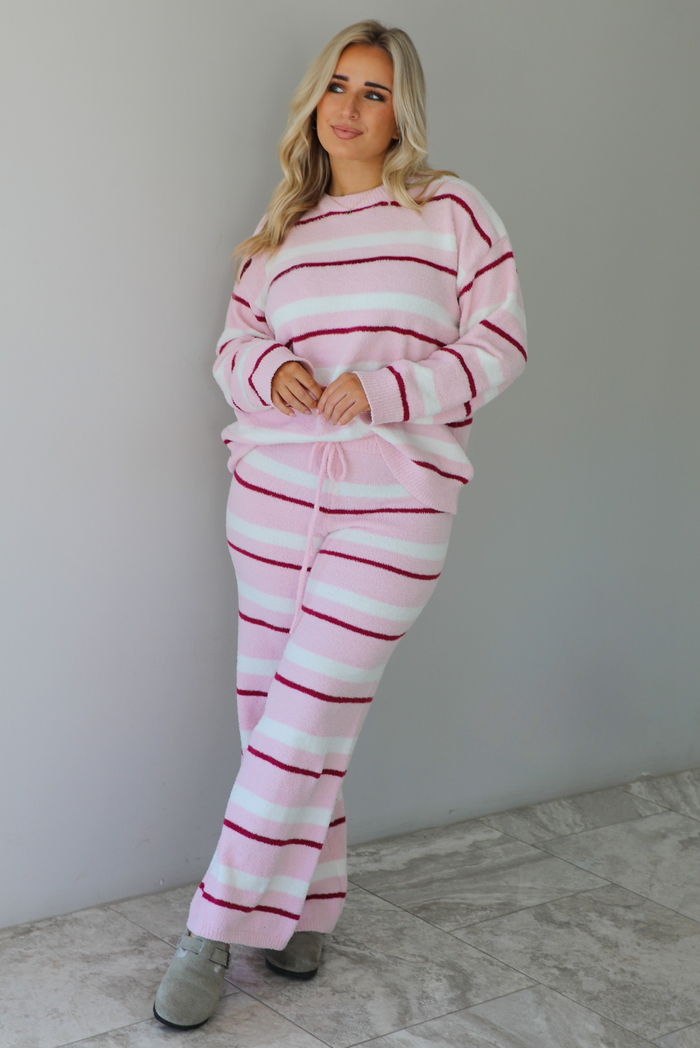 Woman wearing a pink set with a darker pink and white striped design. The set consists of an oversized sweater top and high waisted pants. The photo is taken indoors against a white wall with white marble flooring. 
