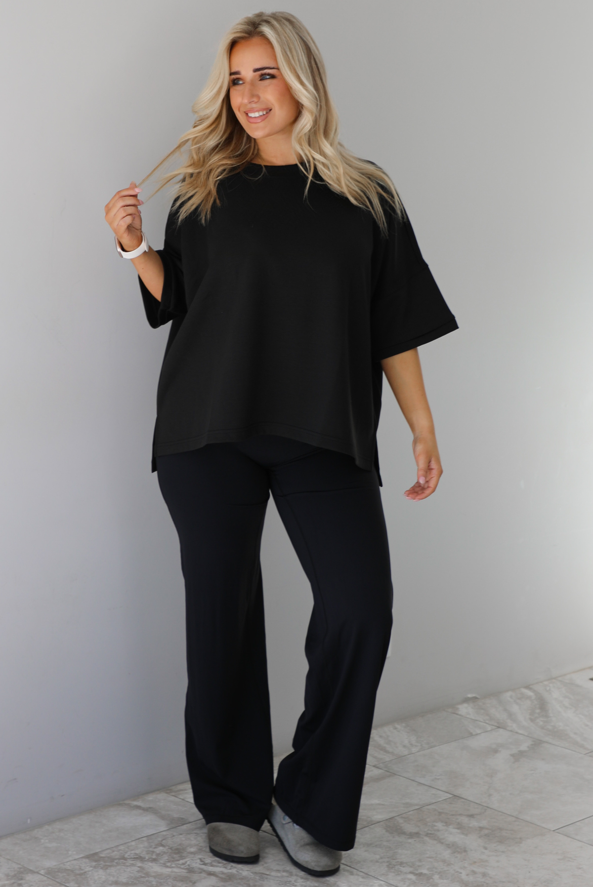 Woman wearing a black oversized box tee with longer short sleeves, a high-low design, and split hem details on either side. The tee is paired with black flared leggings. The photo is taken indoors against a white wall with white marble flooring. 