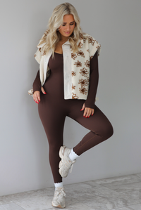 The photo features a model wearing a beige sherpa vest with a brown floral print pattern throughout, functional button closures in the front, and that is oversized with a brown, fitted jumpsuit that's paired with white scrunchie socks and beige sneakers. The photo is taken indoors against a white wall with white marbled flooring. 