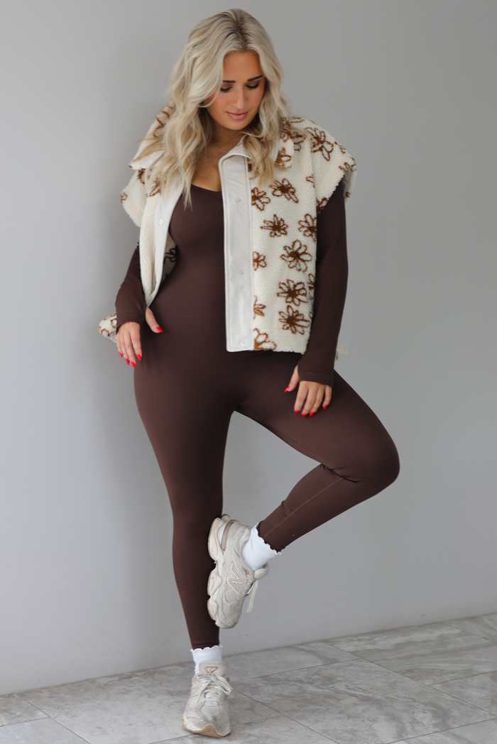 The photo features a model wearing a beige sherpa vest with a brown floral print pattern throughout, functional button closures in the front, and that is oversized with a brown, fitted jumpsuit that's paired with white scrunchie socks and beige sneakers. The photo is taken indoors against a white wall with white marbled flooring. 