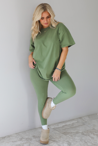 Woman wearing a green two piece set featuring an oversized t-shirt and fitted leggings. The set is paired with high white ankle socks and sand colored warm boots. The photo is taken indoors against a white wall with white marble flooring. 