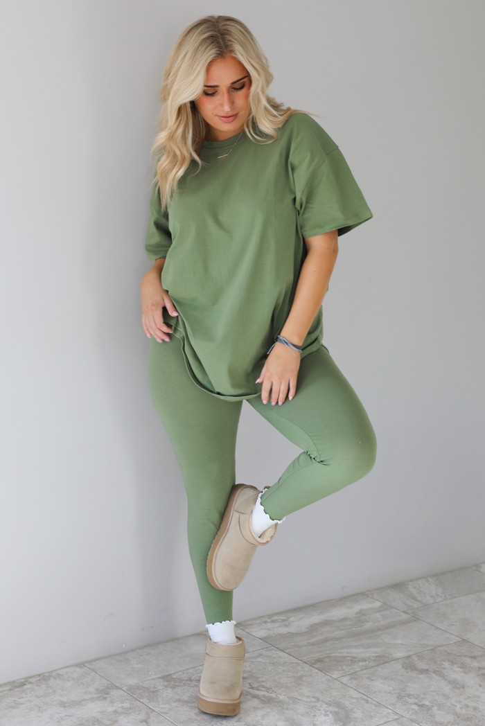 Woman wearing a green two piece set featuring an oversized t-shirt and fitted leggings. The set is paired with high white ankle socks and sand colored warm boots. The photo is taken indoors against a white wall with white marble flooring. 
