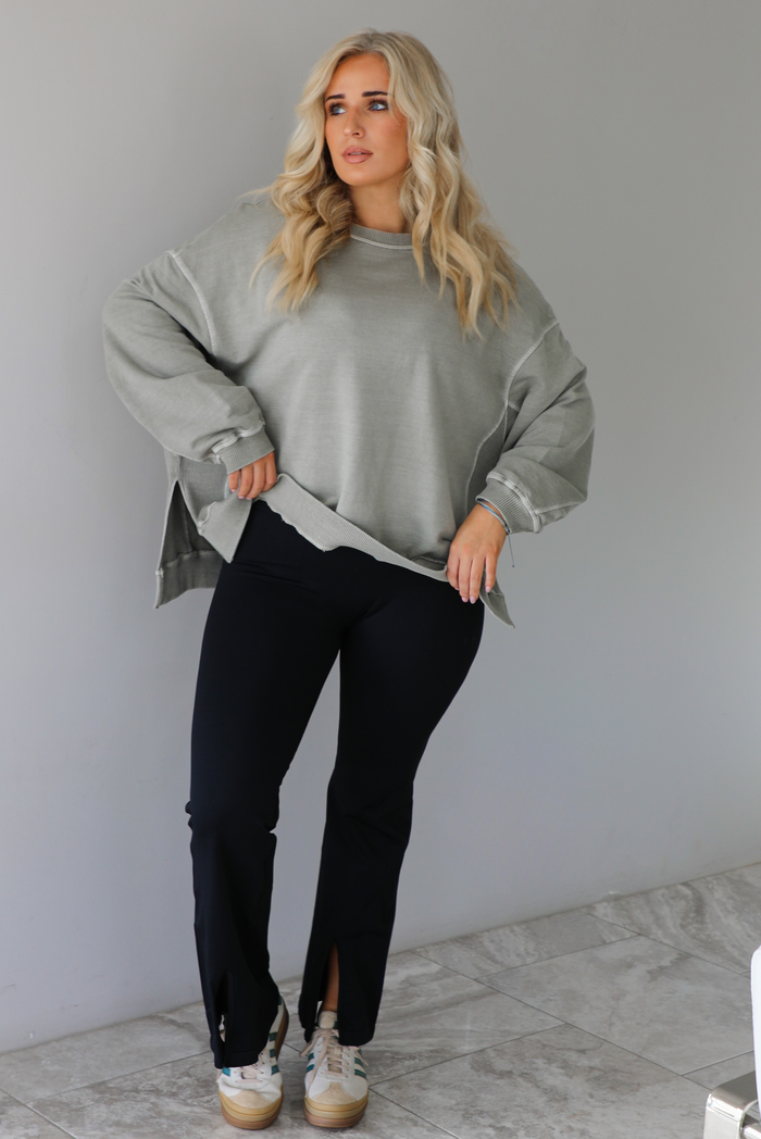 Woman wearing a tan oversized sweater with black leggings that have a slightly flared hem with split hem details in the front. The photo is taken indoors against a white wall with white marble flooring. 