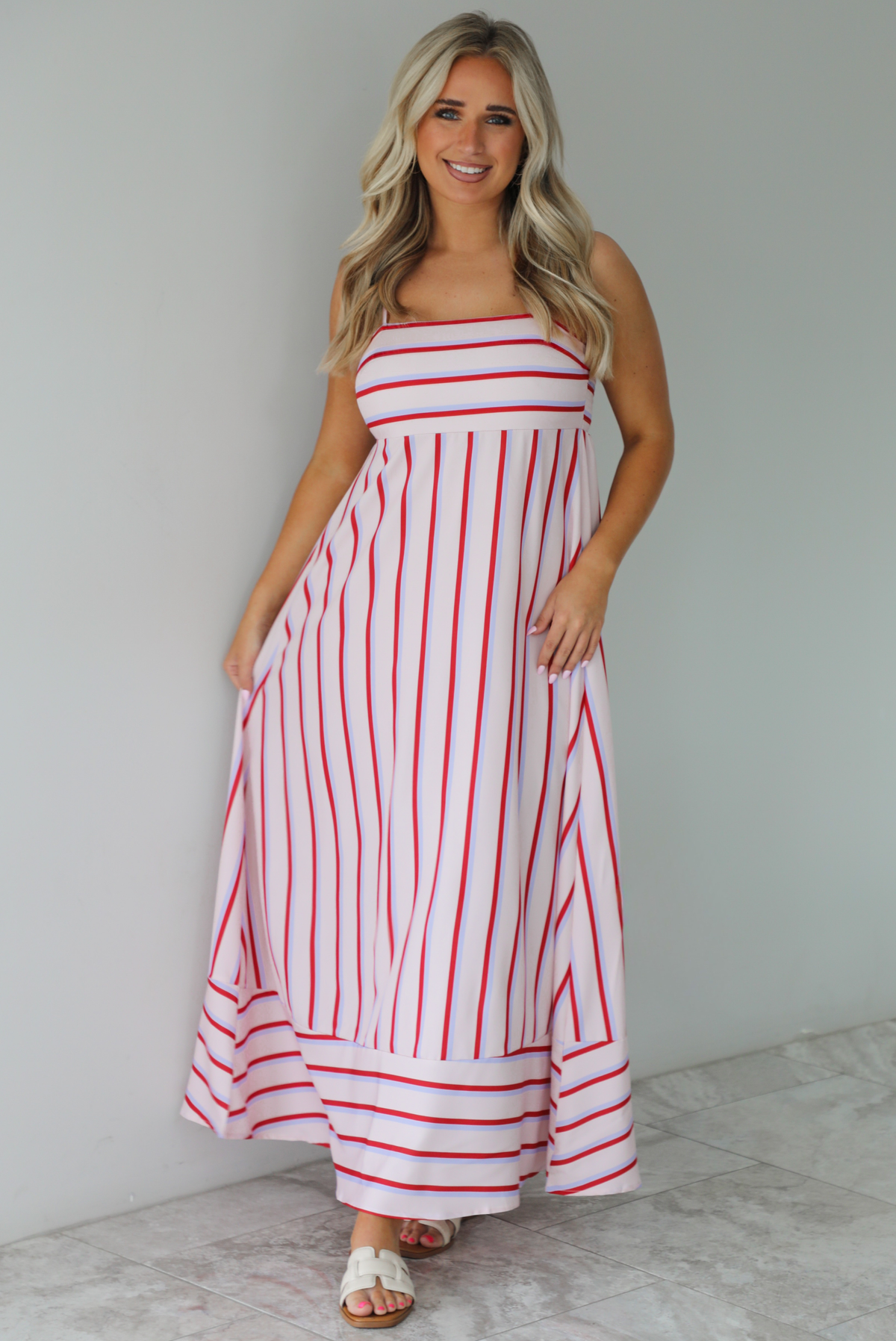 Woman wearing a spaghetti strap, maxi length dress that has a light pink color throughout with a red and light blue striped pattern. The photo is taken indoors against a white wall with white marble flooring. 