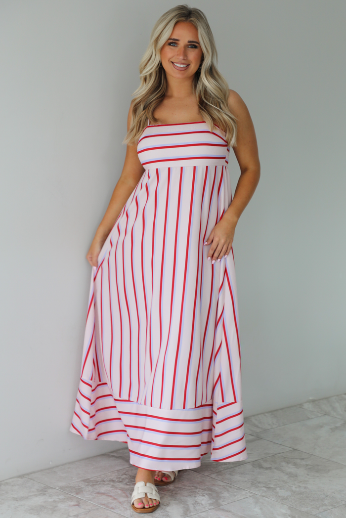 Woman wearing a spaghetti strap, maxi length dress that has a light pink color throughout with a red and light blue striped pattern. The photo is taken indoors against a white wall with white marble flooring. 