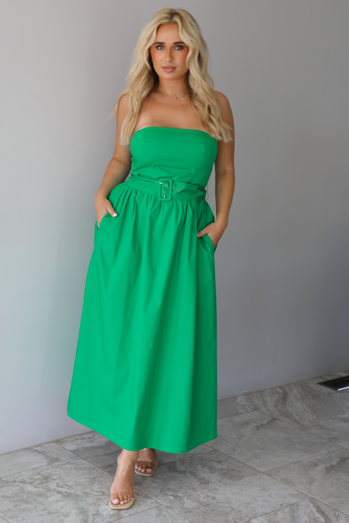 Woman wearing a green strapless midi dress that has a fitted bodice, belted waistline detail, functional pockets on either side, and a midi length indoors against a white wall with white marble flooring. 