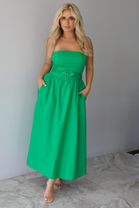 Woman wearing a green strapless midi dress that has a fitted bodice, belted waistline detail, functional pockets on either side, and a midi length indoors against a white wall with white marble flooring. 