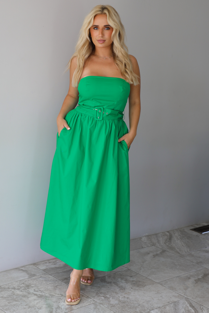 Woman wearing a green strapless midi dress that has a fitted bodice, belted waistline detail, functional pockets on either side, and a midi length indoors against a white wall with white marble flooring. 
