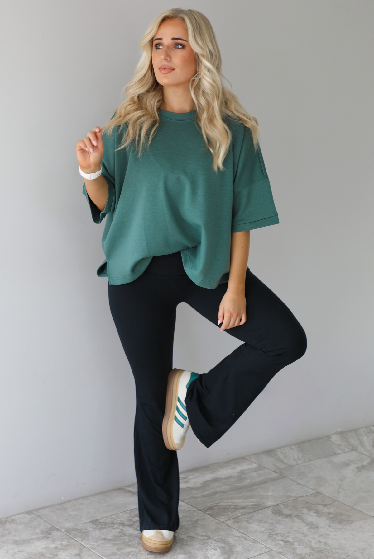 Woman in a teal oversized t-shirt paired with black leggings that flare slightly at the hem. The photo is taken indoors against a white wall with white marble flooring. 