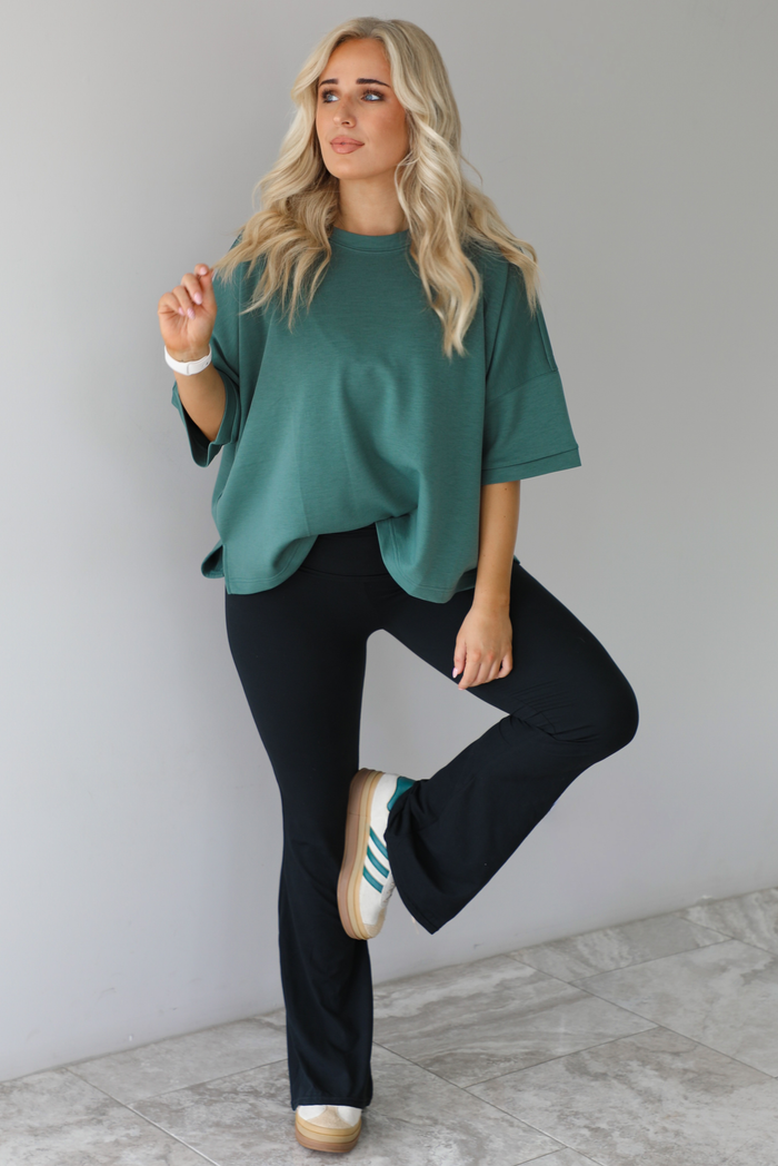 Woman in a teal oversized t-shirt paired with black leggings that flare slightly at the hem. The photo is taken indoors against a white wall with white marble flooring. 