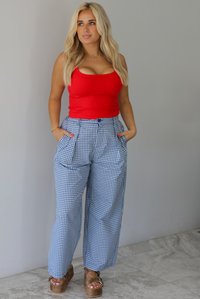 Woman wearing a red tank top high waisted navy and white plaid pants indoors against a white wall with white marble flooring. 