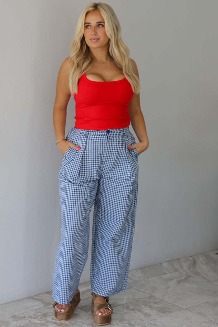 Woman wearing a red tank top high waisted navy and white plaid pants indoors against a white wall with white marble flooring. 
