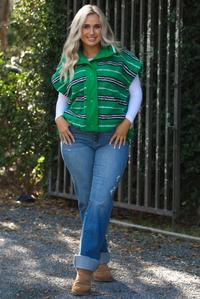 Woman model wearing a green, black, and white striped vest that has a sherpa texture to it, functional button closures in the front, and tie closures on either side, It is paired with a white long sleeved top, a medium wash pair of denim jeans, and tan ugg boots. The photo is taken outdoors with a metal gate and greenery in the back.