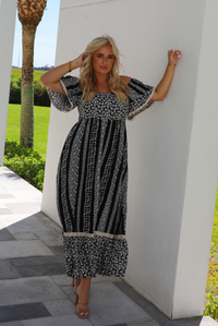 A woman wearing a black and white maxi dress with a white floral print that alternates between a cluster appearance and a striped appearance, with off the shoulder sleeves and a flowy skirt, standing outdoors against a white wall on a stone path with greenery in the background. 