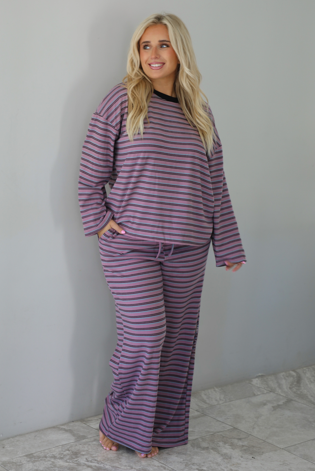 A woman wearing a purple matching set with a black and blue striped pattern featuring an oversized long sleeved top and high waisted flared hem pants, standing indoors against a white wall with white marble flooring. 