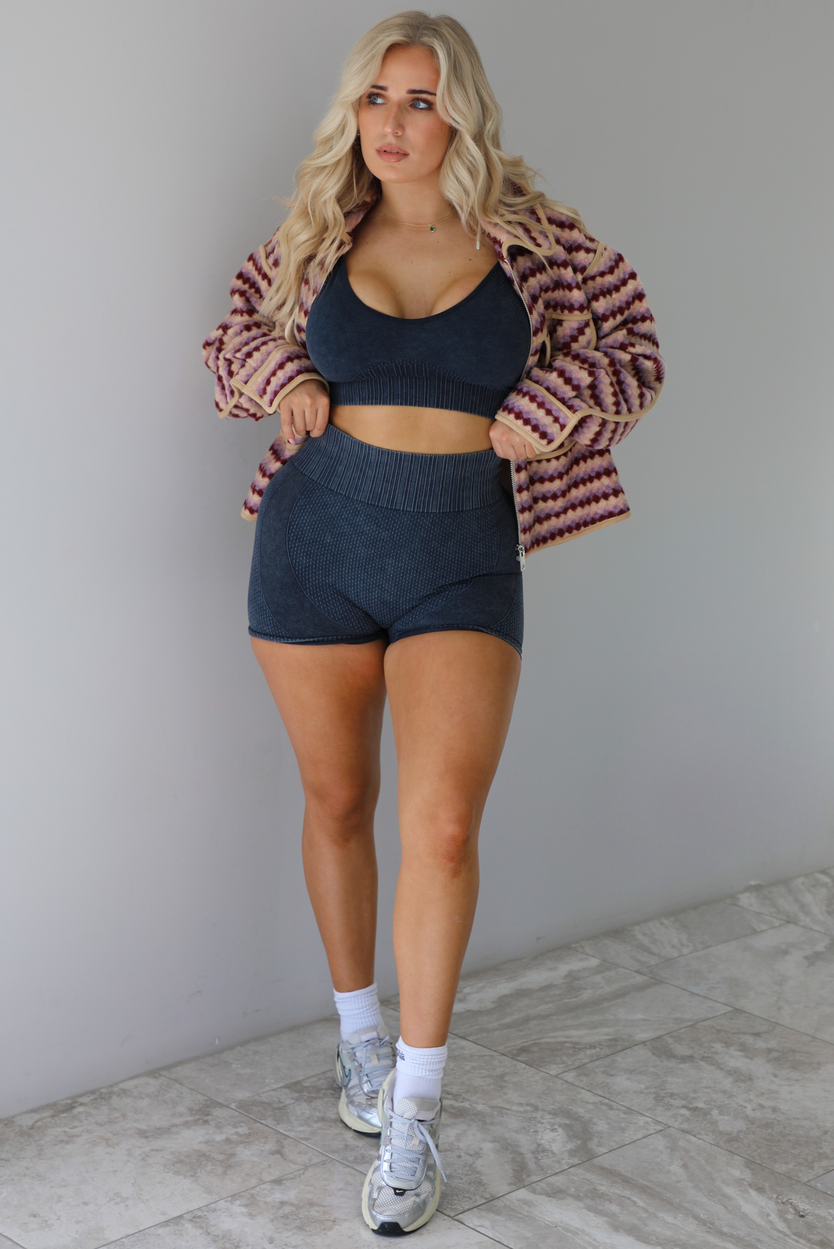 Woman wearing a pink, burgundy, lilac, and sand colored jacket with a charcoal colored two-piece set featuring a cropped sports bra and high waisted short biker shorts. The photo is taken indoors against a white wall with white marble flooring. 