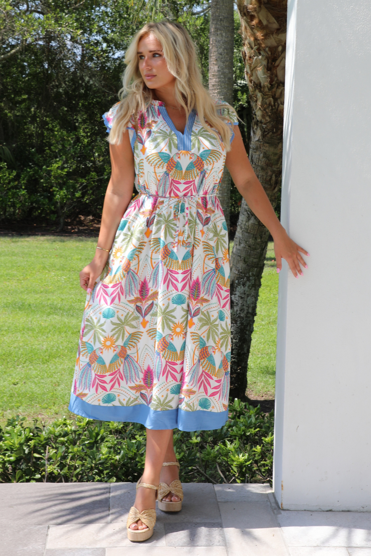 A woman wearing a short sleeved midi dress with a colorful tropical leaf print and light blue trim, standing outdoors next to a white wall with greenery and palm trees in the background. 