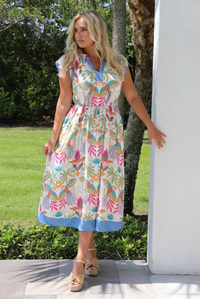 A woman wearing a short sleeved midi dress with a colorful tropical leaf print and light blue trim, standing outdoors next to a white wall with greenery and palm trees in the background. 