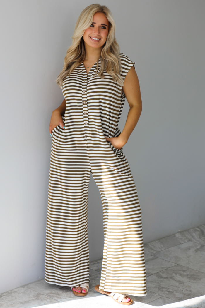 Mocha and white striped patterned jumpsuit that has functional button closures along the front and functional pockets on either side with flared hem bottoms. The photo is taken indoors against a white wall with white marble flooring. 