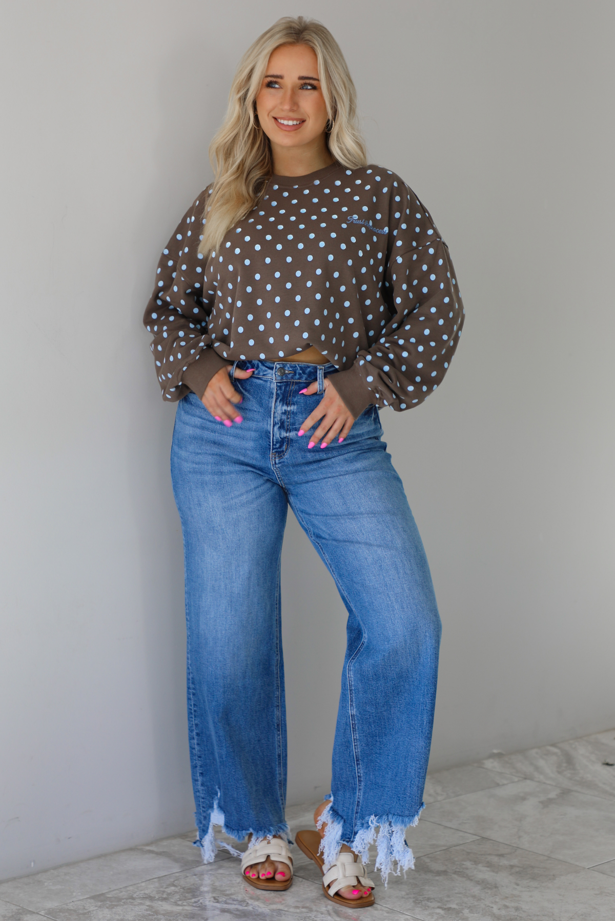 Woman wearing a brown sweater that has a blue polka dot print pattern throughout with blue embroidered writing on the left side that reads 'Trust The Process.' The sweater is paired with a medium wash denim. The photo is taken indoors against a white wall with white marble flooring.