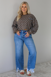 Woman wearing a brown sweater that has a blue polka dot print pattern throughout with blue embroidered writing on the left side that reads 'Trust The Process.' The sweater is paired with a medium wash denim. The photo is taken indoors against a white wall with white marble flooring.