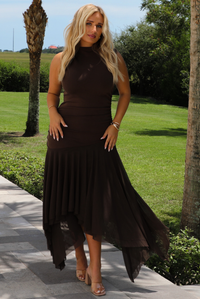 A woman wearing a brown midi dress that's fitted until the upper thigh then leads to sheer mesh that's flowy ands an asymmetrical hem, standing outdoors on a stone path with palm trees and other greenery in the background. 