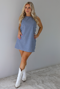 Woman wearing a navy and white gingham print patterned dress that has non-functional button closures on the left side. The photo is taken indoors against a white wall with white marble flooring.