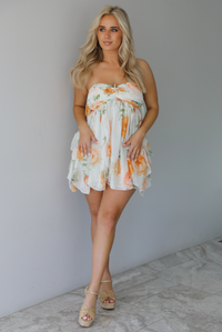 Woman wearing a white, strapless, babydoll cut dress that has an orange and green floral print pattern and flowy skirt indoors against a white wall with white marble flooring. 