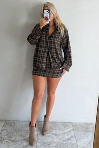 A woman taking a mirror selfie wearing aa dark brown plaid set featuring an oversized button-down and fitted mini length skirt, standing indoors against a white wall with white marble flooring. 