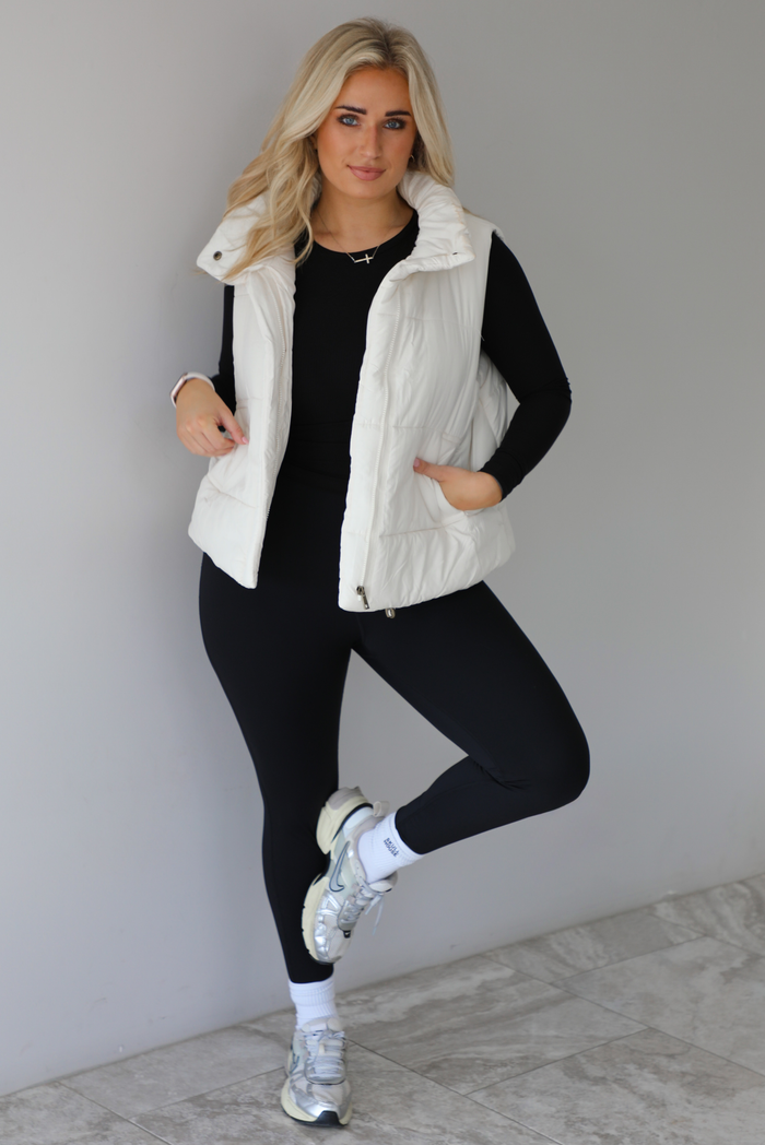 Woman wearing a light sand colored puffer vest with a fitted black long sleeved top and fitted leggings with high white ankle socks and silver sneakers. The photo is taken indoors against a white wall with white marble flooring. 