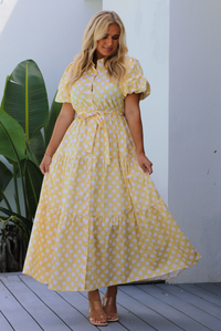A woman wearing a yellow and white polka dot midi dress with puff sleeves and a tie waist and functional button closures along the front, standing on a gray wooden deck against a white wall with a green plant in the background.