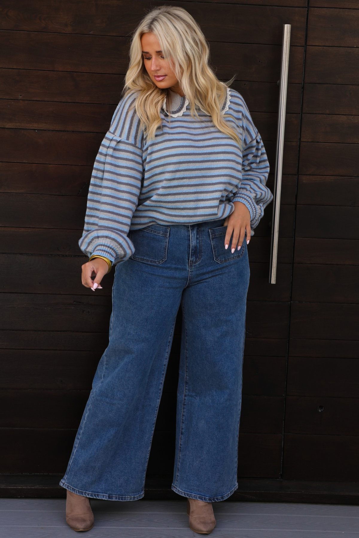 A woman wearing a blue and navy striped long-sleeve sweater that has a bib detail at the neckline and wide-leg blue jeans, standing in front of a dark wood-paneled door.