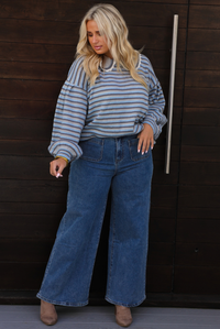 A woman wearing a blue and navy striped long-sleeve sweater that has a bib detail at the neckline and wide-leg blue jeans, standing in front of a dark wood-paneled door.