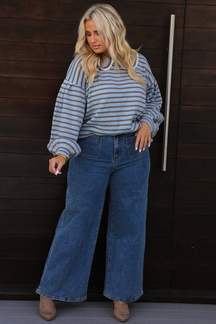 A woman wearing a blue and navy striped long-sleeve sweater that has a bib detail at the neckline and wide-leg blue jeans, standing in front of a dark wood-paneled door.