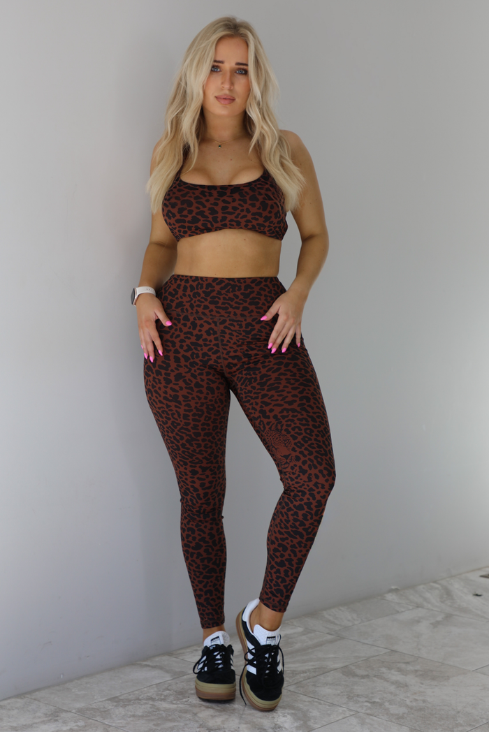 Woman wearing a brown and white leopard print two piece athleisure set that consists of a sports bra and high waisted leggings. The photo is taken indoors against a white wall with white marble flooring. 