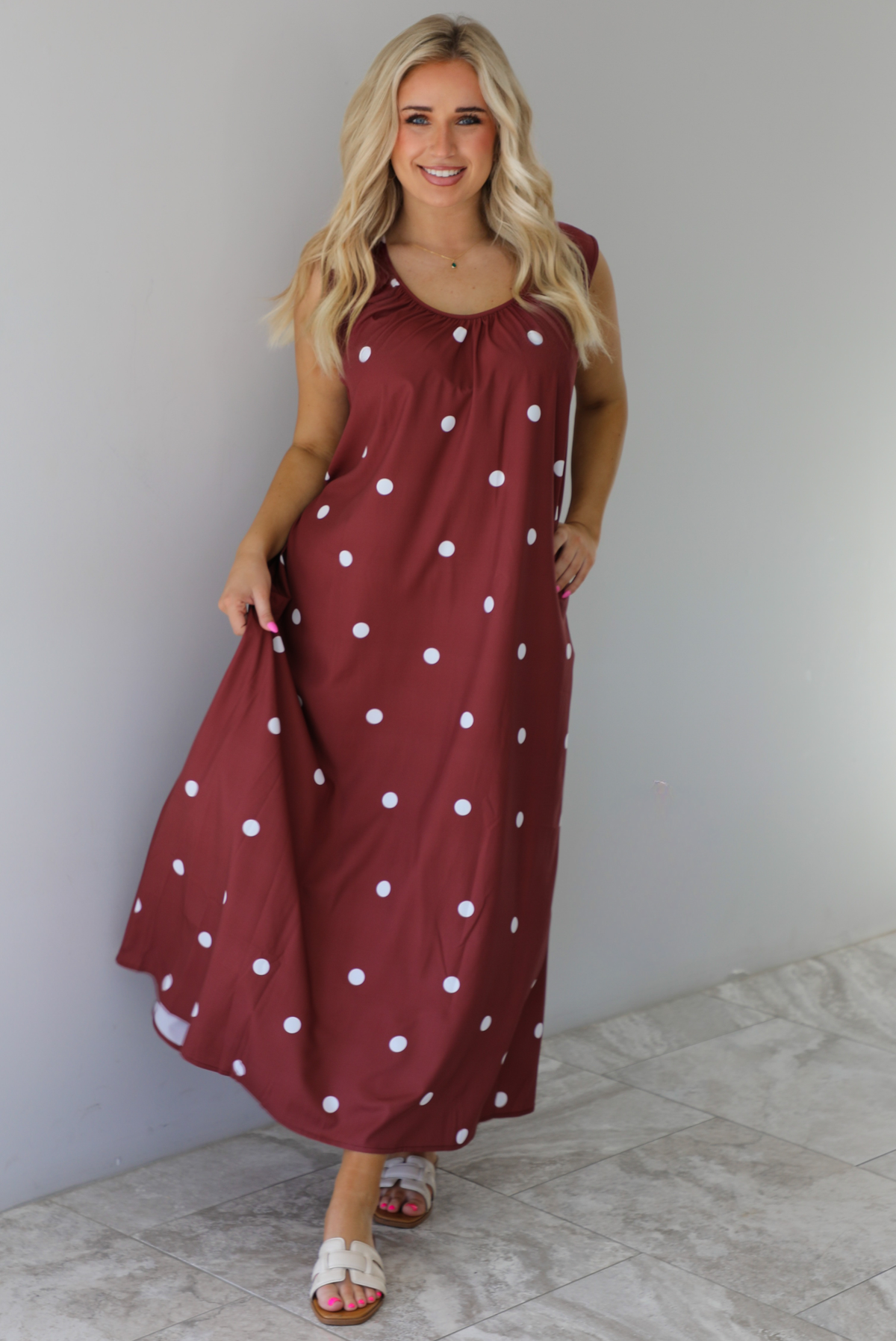 Woman wearing a red brown maxi length dress that has a white polka dot print pattern throughout. The dress is paired with sand sandals. The photo is taken indoors against a white wall with white marble flooring. 