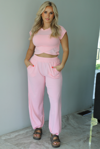 Woman wearing a pink two piece set featuring a cropped short sleeved top and high waisted jogger-style pants indoors against a white wall with white marble flooring. 