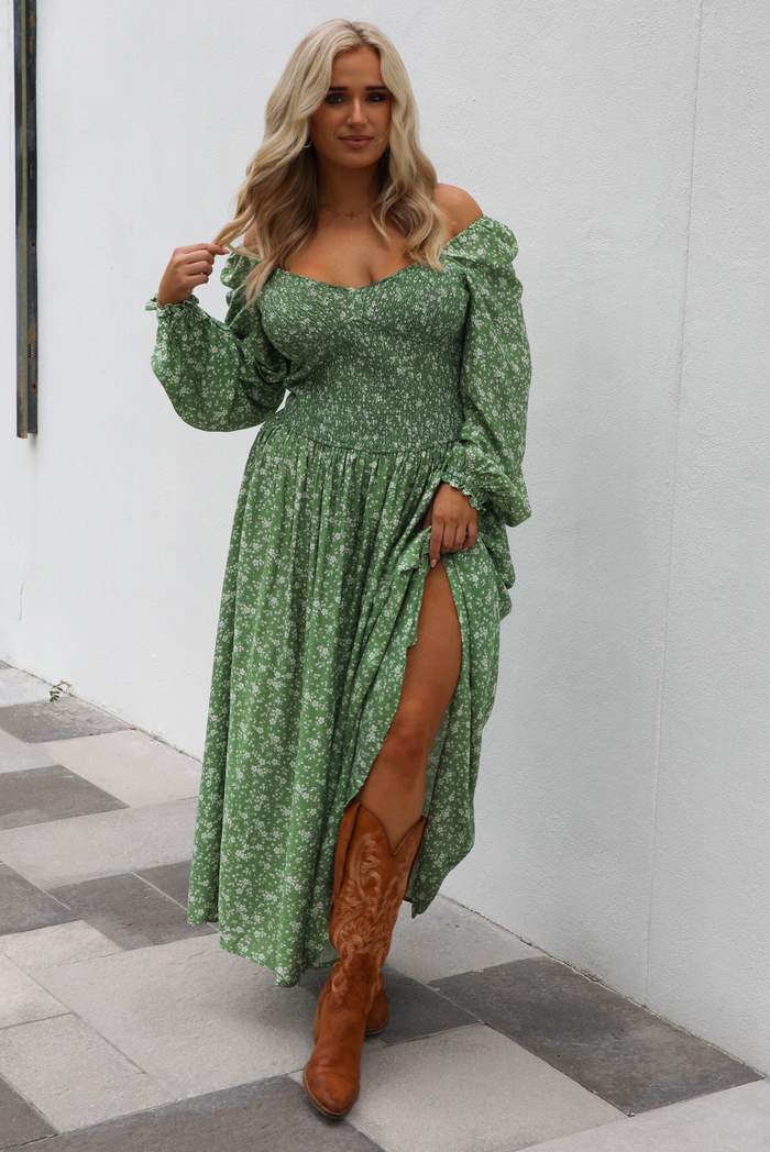 Woman model wearing a green maxi length dress that has a white floral print pattern, long sleeves, smocked bodice, and a flowy skirt. The model is holding the skirt up on one side. The photo is taken outdoors against a white wall. 
